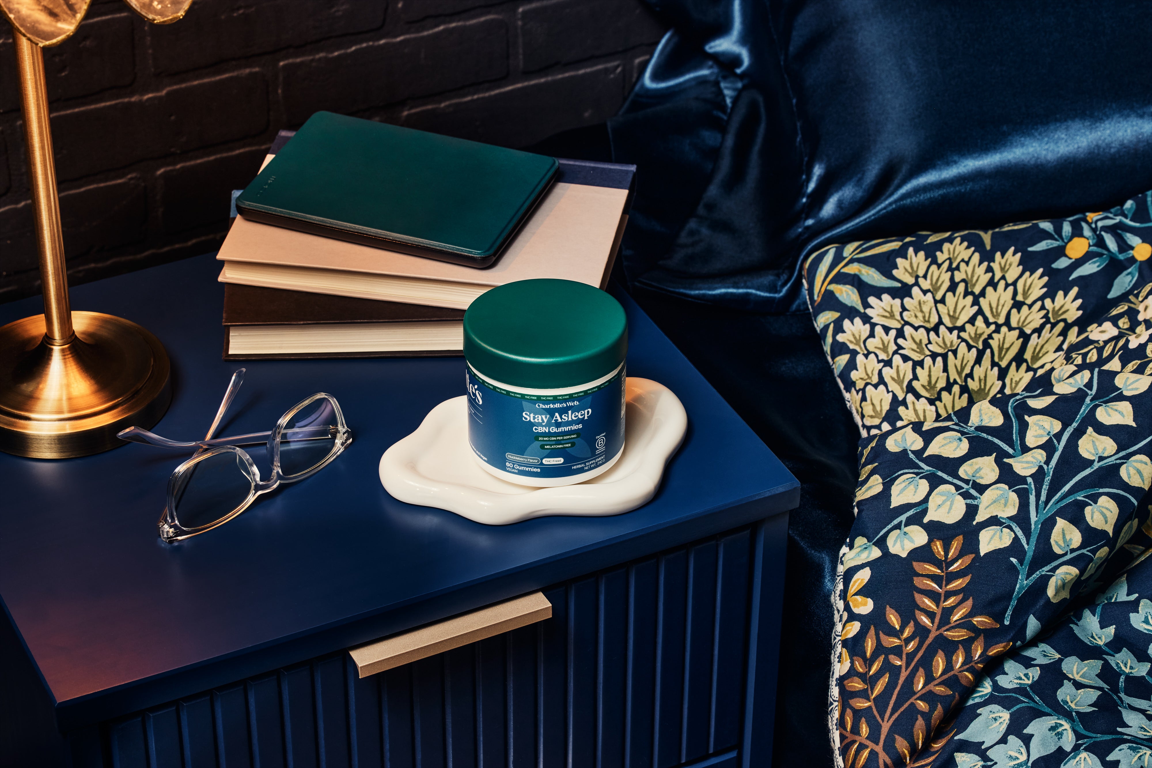 Cosmetic jar on a nightstand with books, glasses, and a lamp in the background.