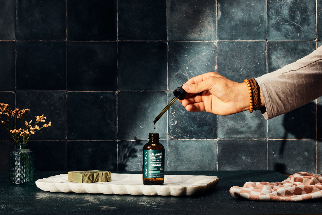 Hand pouring a dropper into a small bottle on a marble tray with a dark tiled wall background