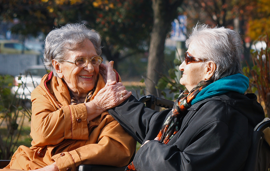 Two elderly women sitting outdoors, one in a wheelchair, with a blurred background