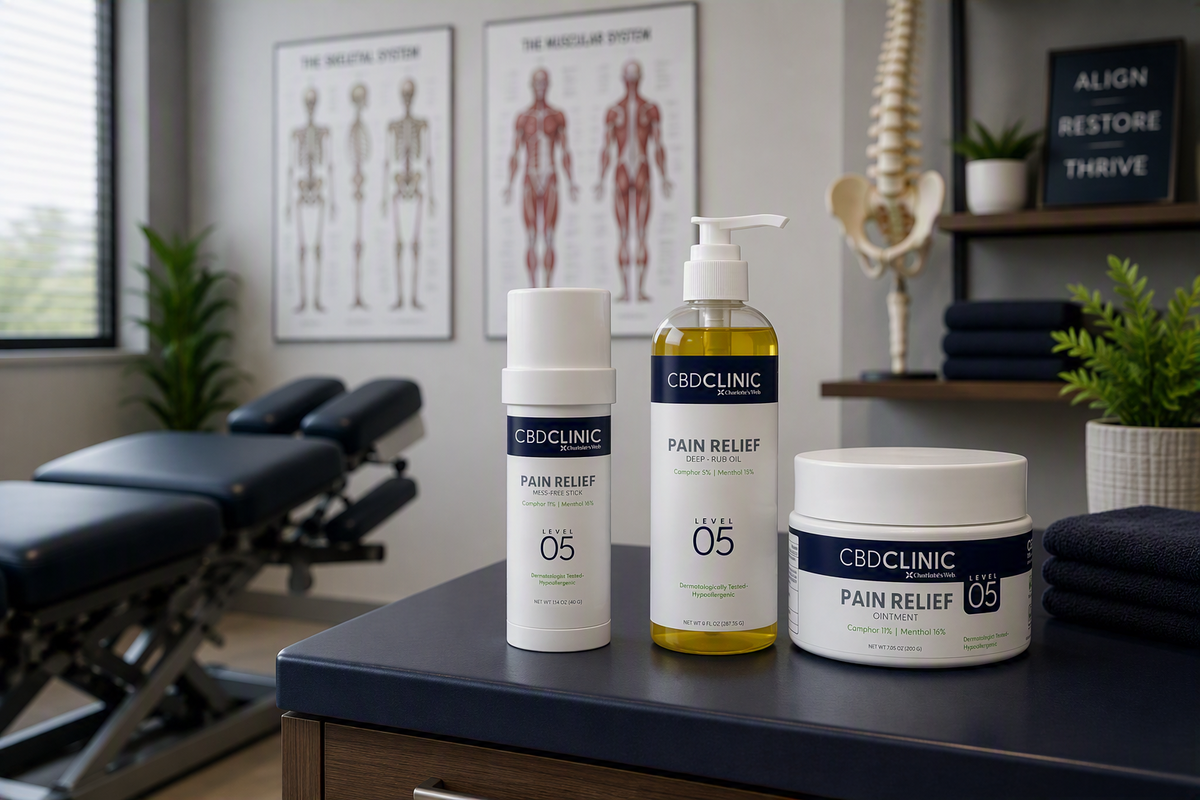 Three CBDCLINIC Pain Relief products (a white cream bottle, a yellow oil pump bottle, and a white cream jar) displayed on a dark blue counter in a treatment room setting.