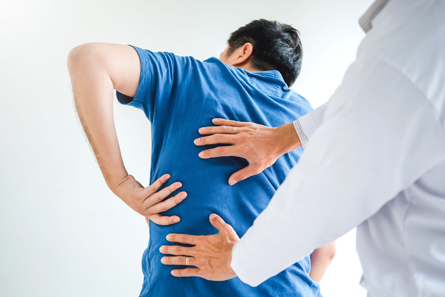 Person in blue shirt holding their back as a doctor examines it.